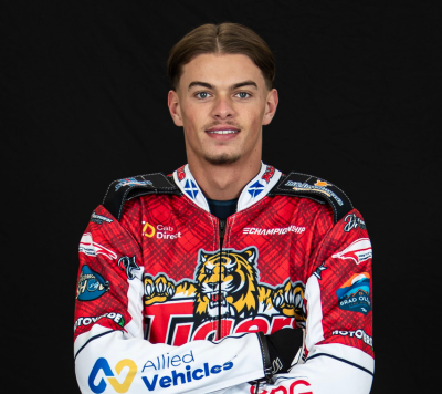 A male speedway rider in a red and white team suit with Tigers and various sponsor logos stands against a black background, facing the camera with arms folded and a neutral expression.