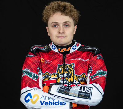 A young speedway rider with curly hair wears a red racing suit featuring sponsor logos and a wildcat graphic, standing against a black background with arms folded.