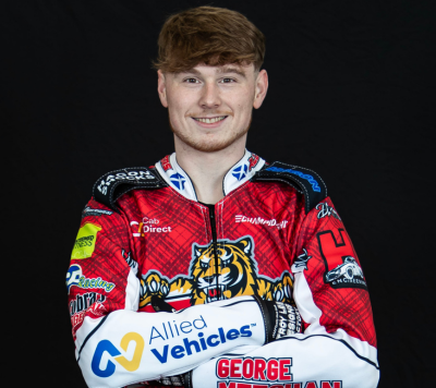 A person in a red racing suit with sponsor logos stands smiling, arms folded, against a black background. The suit features a tiger graphic and the name George is visible.